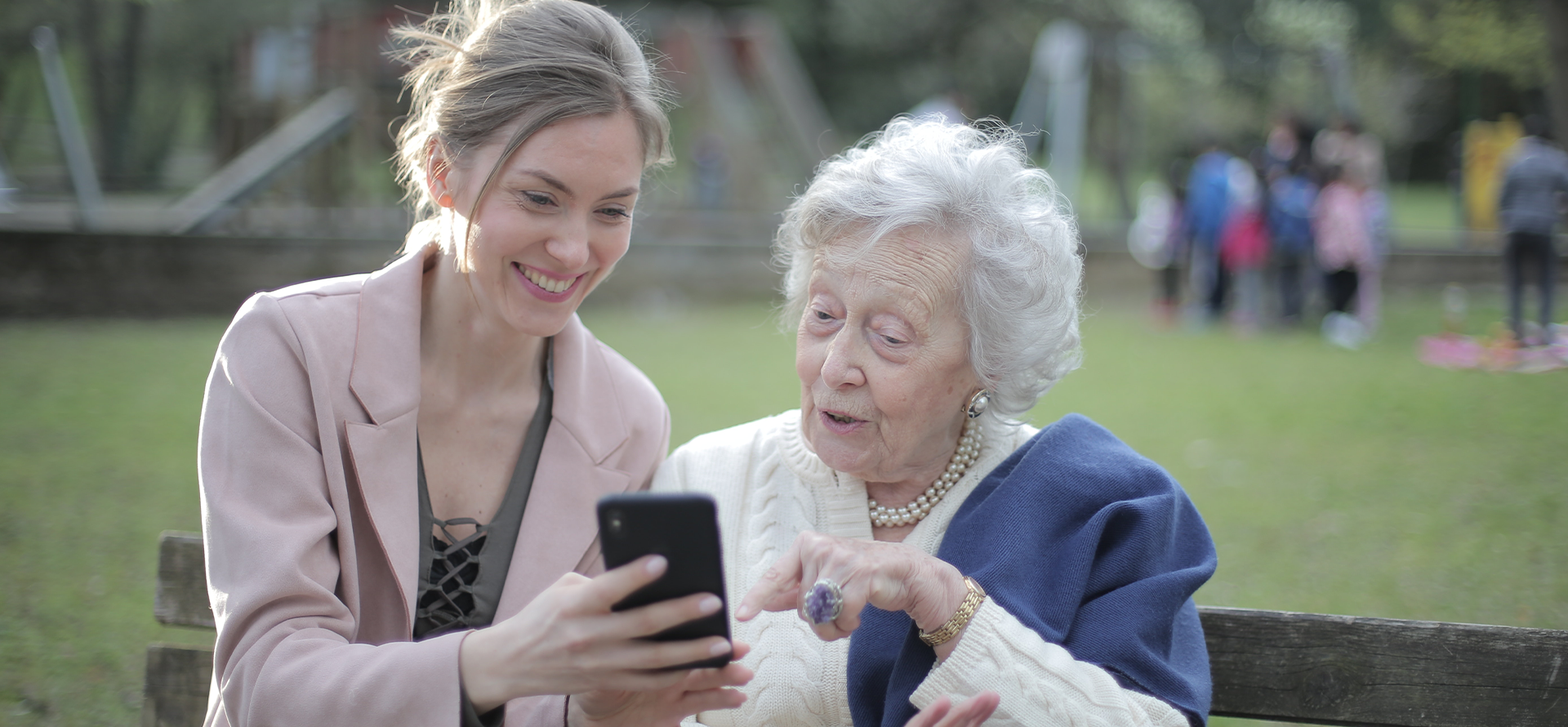 Eine junge Frau und eine Seniorin sitzen zusammen auf einer Bank im Park. Die junge Frau zeigt der älteren etwas auf ihrem Smartphone. Die Seniorin wirkt interessiert.