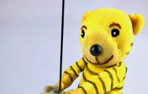 Zusehen ist eine Thaterpuppe der ein gelber Tiger mit schwarzen Streifen sein soll. Der Tiger steht mit beiden Armen nach vorne gerichtet da. Die Wand vor dem der Tiger steht ist Grau.