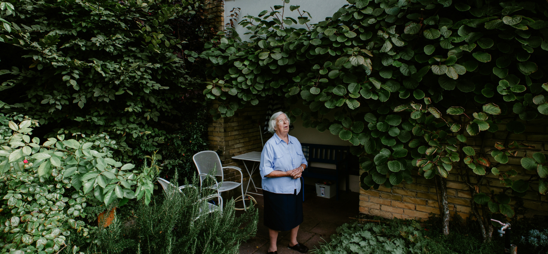 Zusehen ist eine Frau die Nach oben schauend da steht. Im Hintergrund sieht man Zwei weiße Stühle und einen Tisch. Um die Frau herum sind Viele Grüne Pflanzen.