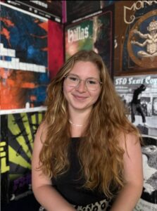 Annika ist unsere FSJ-lerin und unterstützt unsere Arbeit in der Soziokulutr. Sie sitzt mittig und lächelt in die Kamera, vor einem bunten Werbe-Poster-Hintergrund.