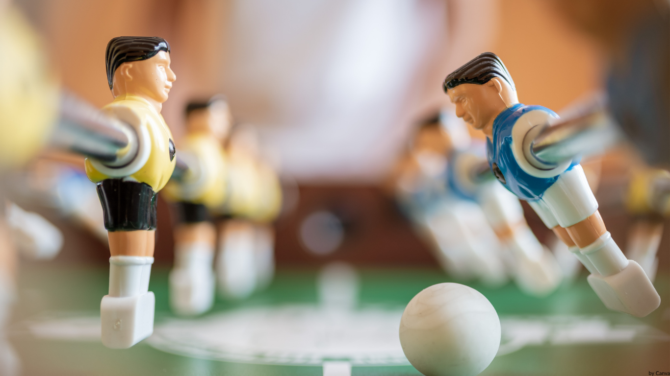 Auf dem Bild sieht man zwei Kickerstangen An dem Kickerfiguren hängen. Die Kickerfigur auf der Rechten seite holt zum Schuss aus während die Kickerfigur auf der Linken seite in Abwehrstellung steht.